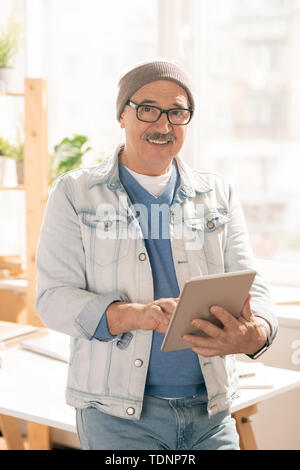 Creative aged man in casualwear looking at you with smile while using digital tablet for searching websites for designers Stock Photo