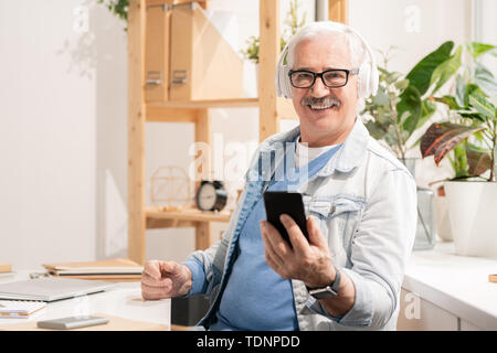Successful aged man in headphones and casualwear using smartphone while sitting by desk in front of camera Stock Photo