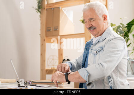 Confident mature man in casualwear pointing at smartwatch on his wrist while checking time before leaving office Stock Photo