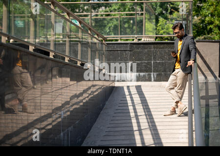Contemporary smart casual businessman standing by railings in urban environment on summer day and texting in smartphone Stock Photo