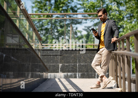 Young casual man in beige jeans, yellow pullover and dark blue jacket scrolling in smartphone by railings outdoors Stock Photo