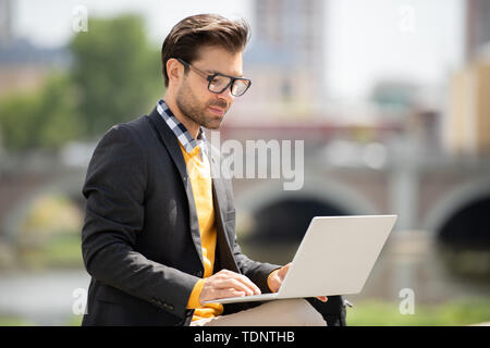 Handsome young freelancer in smart casualwear looking at laptop display while searching for online information outdoors Stock Photo
