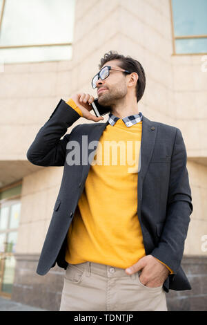 Young elegant man in yellow pullover and dark blue jacket talking on mobile phone against modern architecture Stock Photo