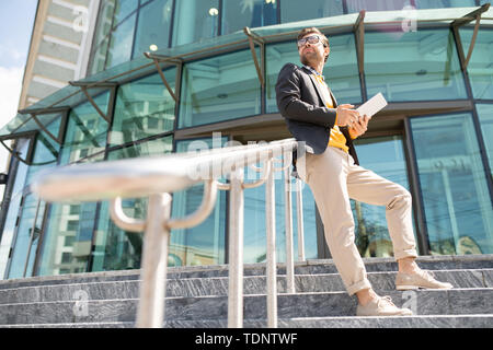 Young contemporary elegant businessman with touchpad standing by banisters on stairs with business center on background Stock Photo