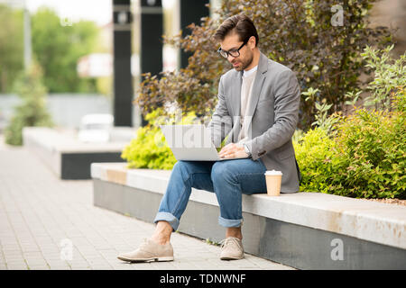 Mobile businessman or freelancer in jacket and jeans sitting in urban environment in front of laptop and working in the net Stock Photo