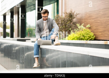 Confident businessman in smart casualwear sitting in front of laptop while networking or video-chatting in urban environment Stock Photo