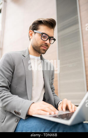 Serious young businessman in smart casual concentrating on working over new project in front of laptop outdoors Stock Photo