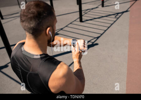 Young contemporary athlete in activewear standing on stadium before running and looking at smartwatch on his wrist Stock Photo