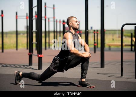 Strong young athlete in activewear doing exercise for stretching legs on outdoor sportsground at leisure Stock Photo