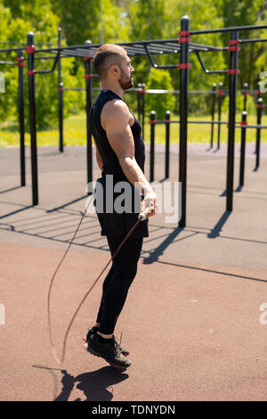 Young muscular athlete in tracksuit jumping with skipping-rope on sunny morning during outdoor workout Stock Photo
