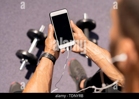 Smartphone in hands of young muscular athlete surfing in playlist while listening to music at break Stock Photo