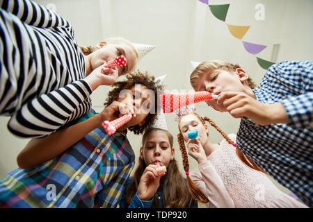 Low angle view at group of happy kids blowing party horns at camera and having fun Stock Photo