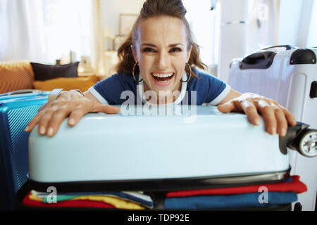 Portrait of happy modern woman in blue t-shirt in the modern house in sunny summer day packing suitcase. Stock Photo