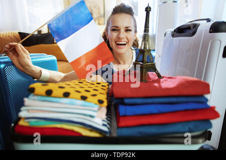 happy stylish traveller woman in blue t-shirt at modern home in sunny summer day showing French flag and Eiffel tower souvenir. Stock Photo