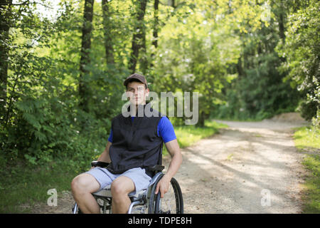 Happy and young disabled or handicapped man sitting on a wheelchair in nature turning wheels on a walking road at a beautiful sunny day Stock Photo