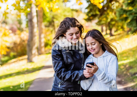 Two sisters looking at social media on a cell phone while walking down a path  during a family outing in a city park on a warm autumn day: Edmonton... Stock Photo