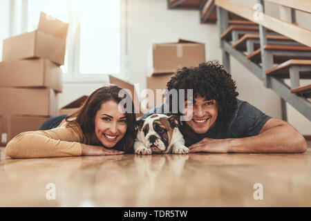 The young happy couple is moving into a new house. They are lying down on the floor and hugging their little puppy after they brought boxes with thing Stock Photo