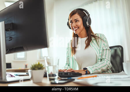 A successful female entrepreneur, with headphones on her head, is listening music on a smartphone and  working on the computer in the office. Stock Photo