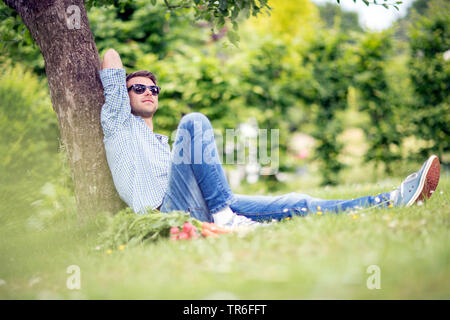 lying with fresh vegetables on lawn in a garden, leaning against a tree trunk, Germany Stock Photo