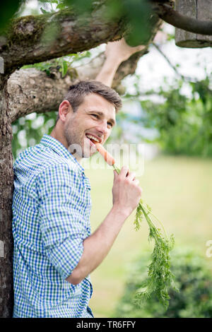 young man biting in freshly harvested carrot in a garden, Germany Stock Photo