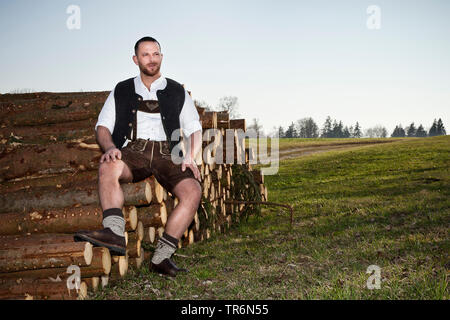 man in Bavarian traditional clothing sitting on felled tree trunks, Germany, Bavaria Stock Photo