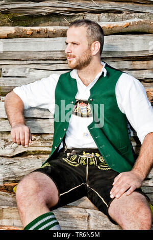 man in Bavarian traditional clothing sitting on a wooden pile, Germany, Bavaria Stock Photo