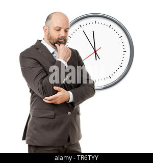 Middle-aged businessmann reflecting with hand touching his chin, clock in the background, Germany Stock Photo