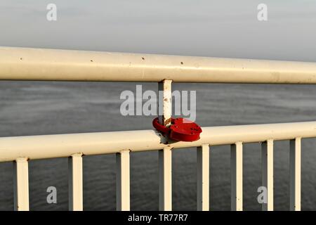 A red metal lock, in the shape of a heart, hangs on the slightly rusty white railing of the river embankment. A railing of a metal pipe divides the ba Stock Photo