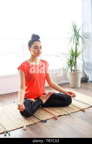 Attractive young brunette woman exercising and sitting in yoga lotus position while resting at home Stock Photo