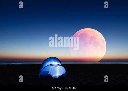 Amazing red moon over the sea, beach and lit tent at sunrise Stock Photo