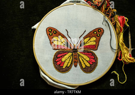 Bright butterfly embroidered on white cotton fabric stretched on a wooden hoop with scissors and colored threads lie on black plywood Stock Photo