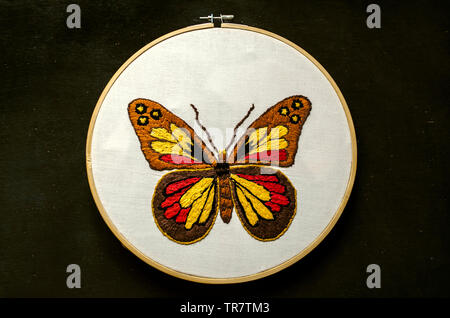 A bright butterfly is embroidered on a white cotton fabric stretched on a wooden Hoop lie on black plywood Stock Photo