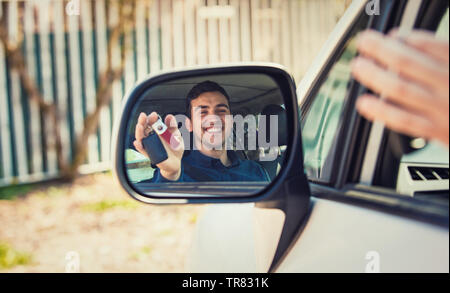 Casual guy driver showing car keys in the side view mirror reflection. Successful young man bought a new car. Rental cars or drivers licence concept, Stock Photo