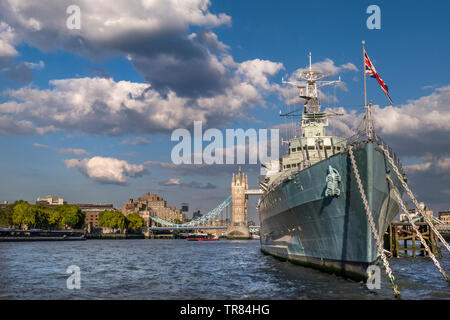 HMS Belfast ship flying Union Jack Flag, moored on River Thames in late afternoon sunlight, with Tower Bridge and The Tower Hotel behind London SE1 Stock Photo