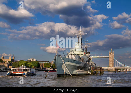 HMS Belfast museum tourist ship moored on River Thames with The Tower of London, a passing RB1 River Clipper Boat and Tower Bridge behind London SE1 Stock Photo