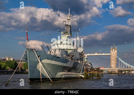 HMS Belfast tourist attraction ship, moored on River Thames in late afternoon sunlight, with Tower Bridge and London Red Bus crossing London SE1 Stock Photo