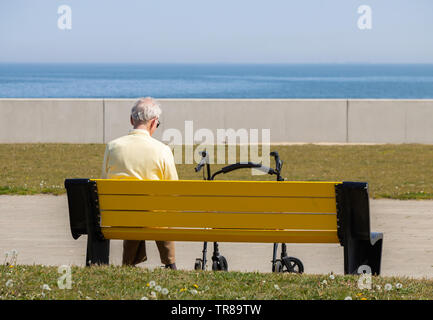 Rear view of elderly man with walker/rollator sitting on seat/bench looking out to sea. UK Stock Photo