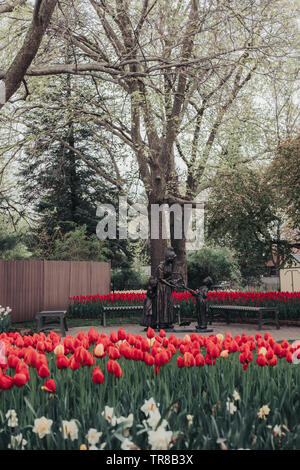 Pella, Iowa, USA - May 2, 2019: Sculpture of a Dutch woman and two children with a basket of tulips in Scholte Gardens. Beds of red and yellow tulips. Stock Photo