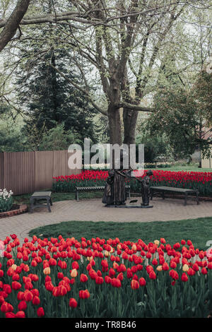 Pella, Iowa, USA - May 2, 2019: Sculpture of a Dutch woman and two children with a basket of tulips in Scholte Gardens. Beds of red and yellow tulips. Stock Photo