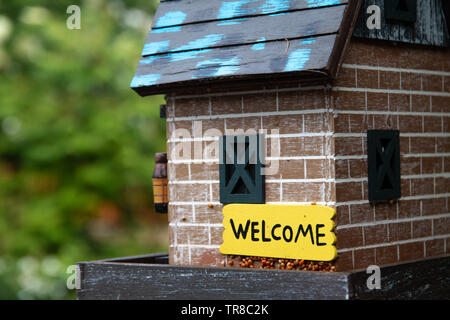 Small bird feeder in old brick barn disguise with some green windows. A welcome sign on the side. Green blurry background. Stock Photo