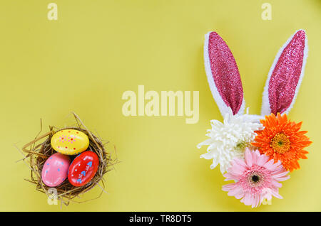 Colorful easter eggs in a nest and easter bunny ear rabbit decoration with gerbera and chrysanthemum flowers on yellow background - top view Stock Photo