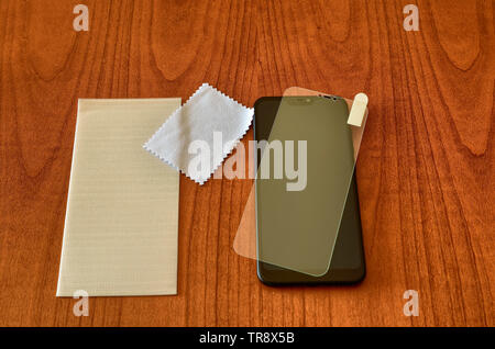 On the wooden table rest the smartphone, the panel and the protective glass to be installed. With a microfibre cloth, detergent residues will be dried Stock Photo