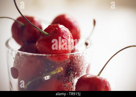 Appetizing ripe red cherry fruits, washed and strewn with droplets of water, lie in a glass vase, illuminated by light. Stock Photo