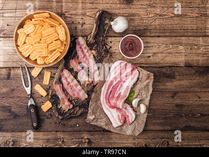 Bacon flavored snacks chips with grilled bacon rashers on vintage chopping board with smoked raw bacon on butchers paper with garlic and sauce on wood Stock Photo