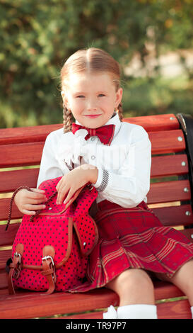 Happy kid girl 5-7 year old sitting on bench with school bag outdoors. Wearing school unifom. Looking at camera. Childhood. Smiling pupil. Stock Photo