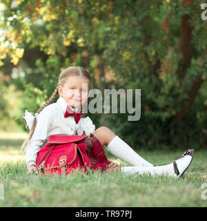 Smiling pupil girl 6-7 year old sitting on grass with backpack outdoors. Looking at camera. Back to school. Childhood. Stock Photo