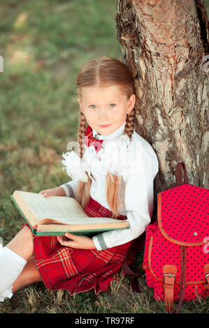 Smiling school girl 6-8 year old reading book outdoors. Wearing stylish school unifom. Looking at camera. Childhood. Back to school. Stock Photo