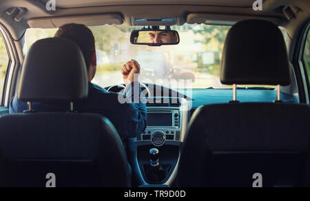 Young man driver sitting in his car behind the wheel shaking his fist threatens another motorist ahead. Bad and aggressive behavior on road concept. N Stock Photo