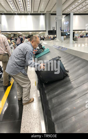 Luggage conveyor belt with personal suitcases moving around circle. Multiracial people waiting for their luggage. Hong Kong International Airport. Stock Photo