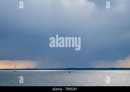 a rainfront moving approaching over Solent with lone single yacht sailing in sea with buoy under big sky storm clouds orange sunset Stock Photo